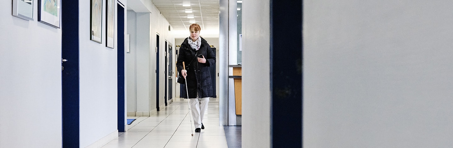 A woman (Sandra Pilz) who is blind using her white cane to guide her through the corridor of BFW Würzburg
